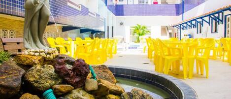 Une cour avec une statue, une fontaine entourée de rochers et des chaises jaunes disposées pour s’asseoir.