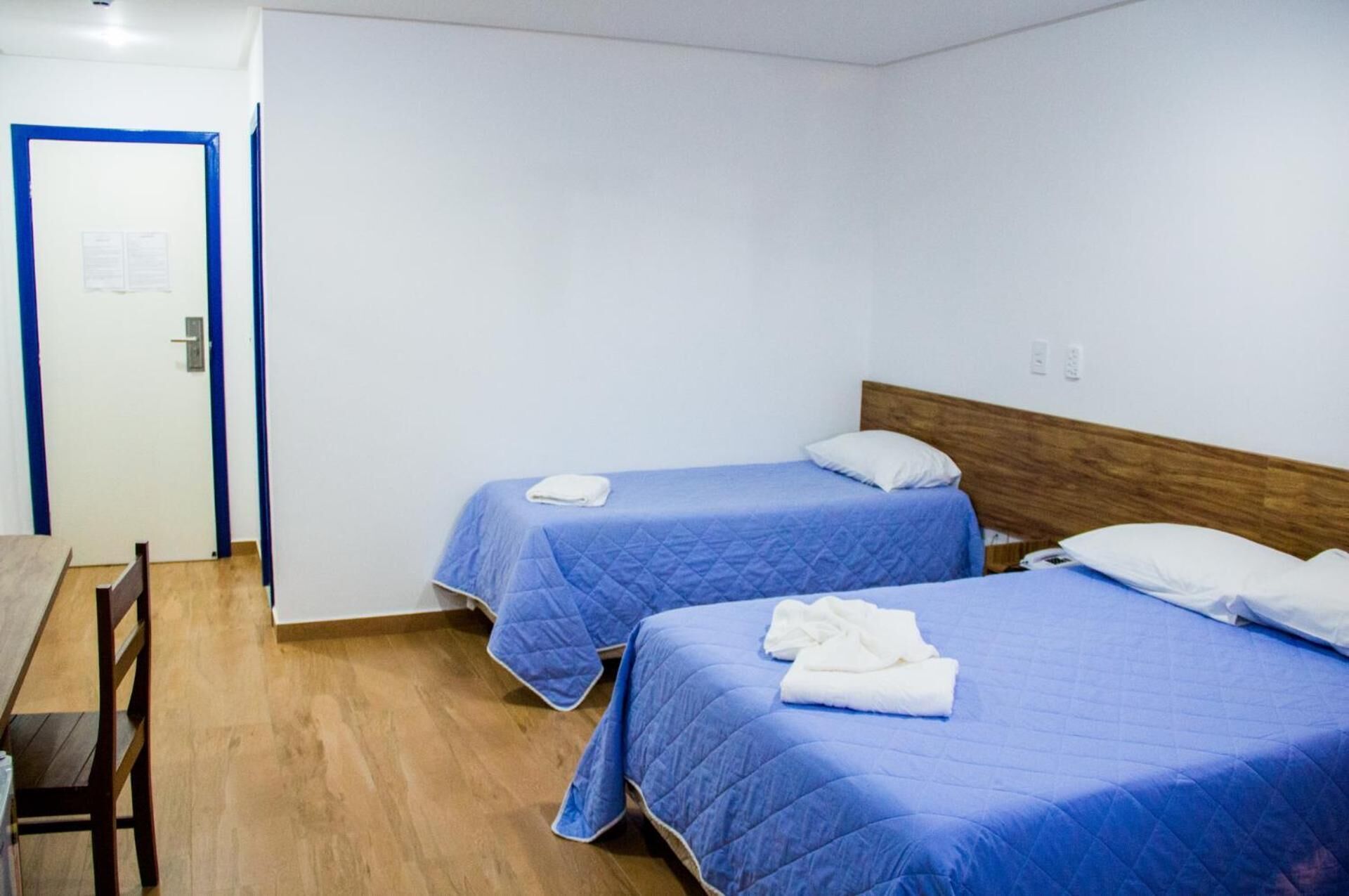 A room with two beds, each with a blue bedspread and white pillows, a wooden headboard, and a wooden table with chairs.