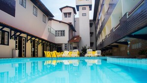 Une piscine entourée de chaises jaunes, un espace de repos couvert et un bâtiment doté de plusieurs balcons.