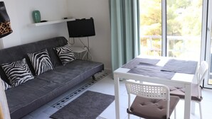 A modern living room with a grey sofa, zebra-patterned pillows, a flat-screen TV on a white shelf, a dining table with a white chair, and a view of trees through large windows. - Cales de Mallorca Studio mit Meerblick / 100m vom Stand / Wifi (Cales de Mallorca)
