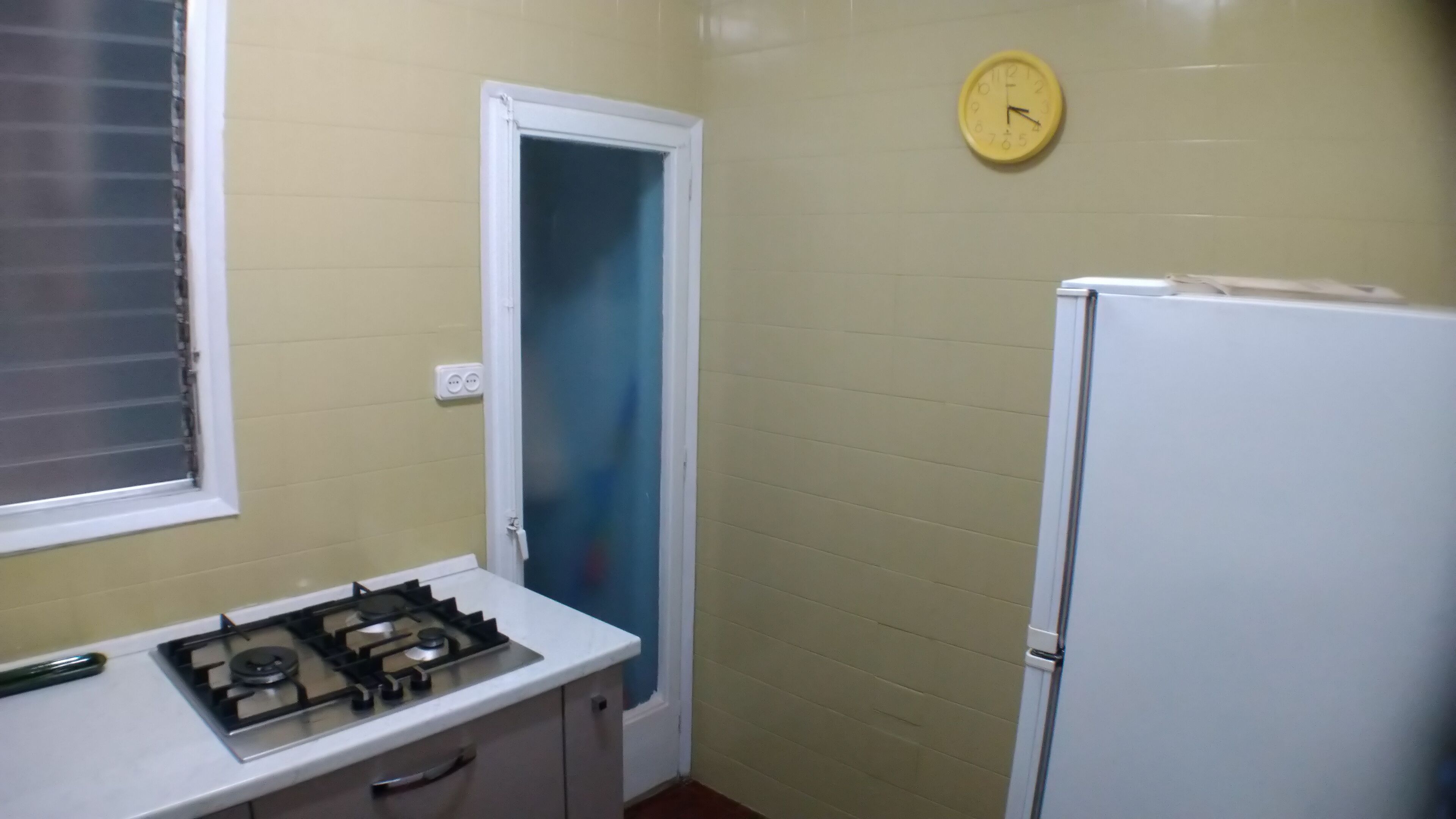 A small kitchen with a gas stove, a white refrigerator, a clock on the wall, and a door leading to another room.