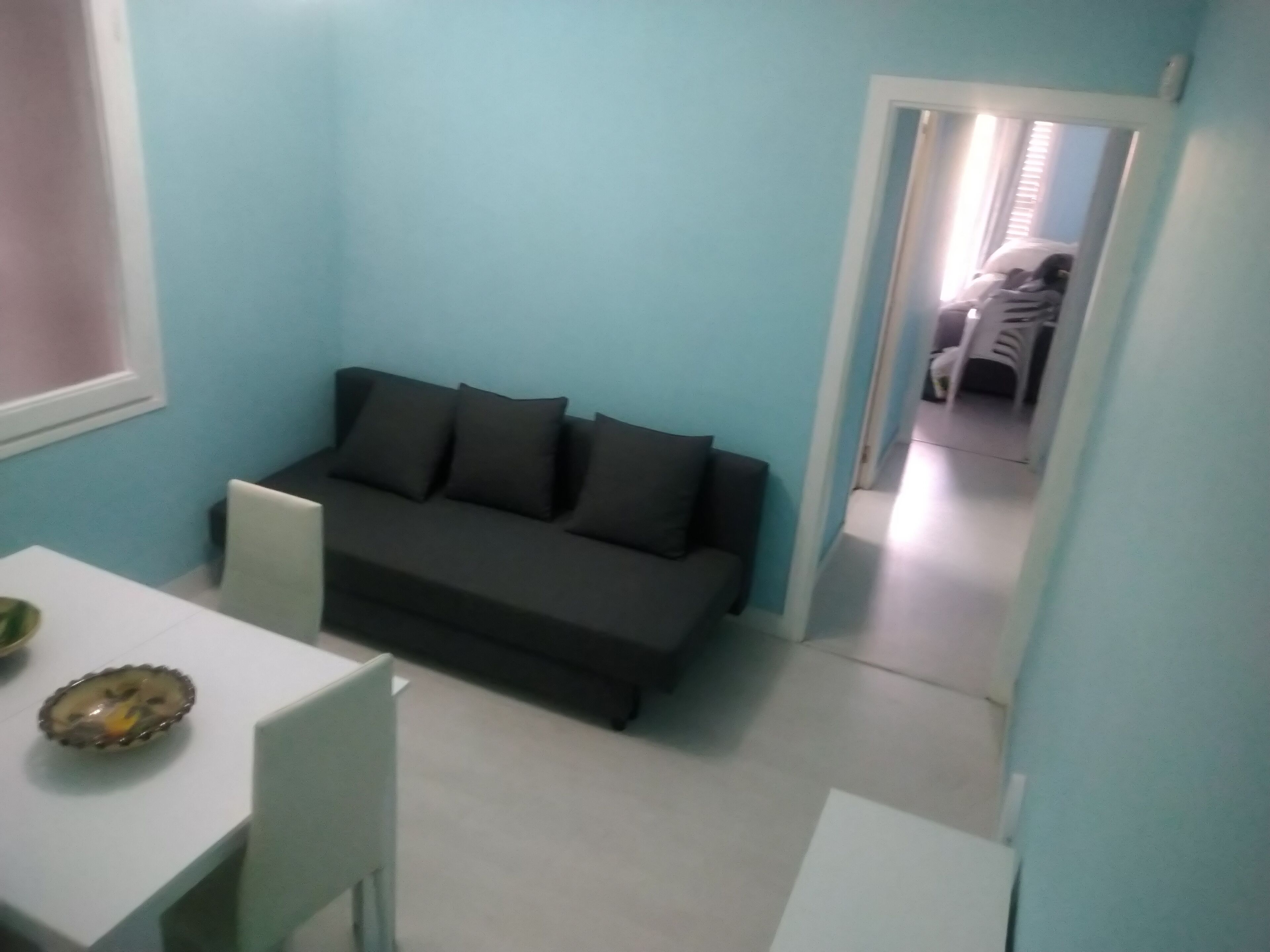 Un espace de vie compact, avec un canapé de couleur sombre placé contre un mur bleu clair, une petite table à manger avec un saladier, et une porte visible donnant sur une autre pièce.