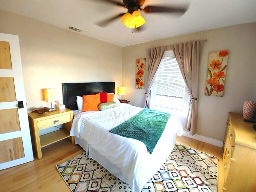 A bedroom with a bed, nightstands, a ceiling fan, a window with curtains, and a door with a wooden panel.