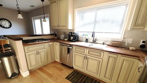 A kitchen with light-colored cabinets, a stainless steel dishwasher, a coffee maker, and a window with blinds.