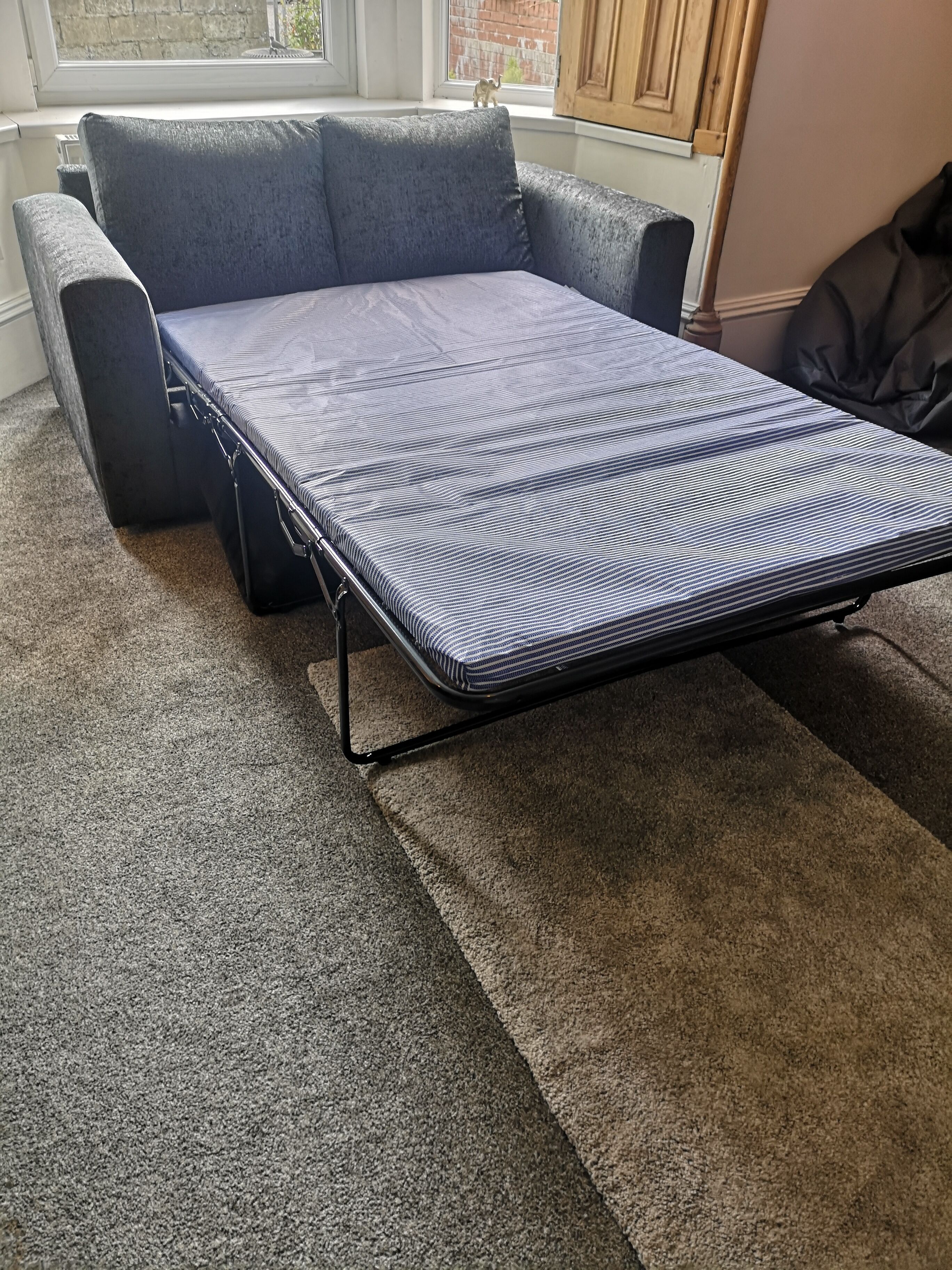 A compact bed in a room with a grey carpet, a grey sofa, and a window with a view of a brick building.