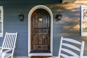 A wooden front door with a white frame, flanked by two white chairs, and two wall-mounted lanterns.
