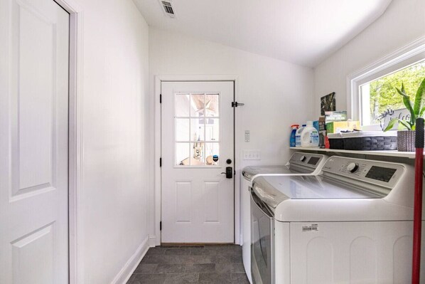 A laundry room with a white door, a washing machine, and a dryer. There is a window with a view of trees outside.