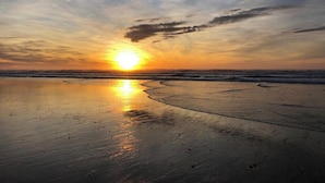 Uma praia ao pôr do sol, com o reflexo do sol na areia molhada.