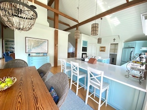 Uma cozinha ampla com uma ilha grande, banquetas brancas e uma mesa de jantar com uma cesta de maçãs.