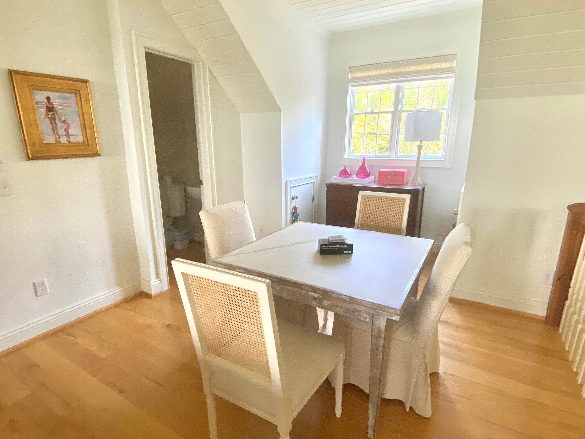 Un coin repas avec une table et des chaises blanches, un tableau encadré au mur et une fenêtre avec des stores.