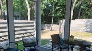 Eine überdachte Terrasse mit Gartenmöbeln, darunter Stühle und ein Sitzsack, mit Blick auf einen Garten mit Holzzaun und Bäumen.