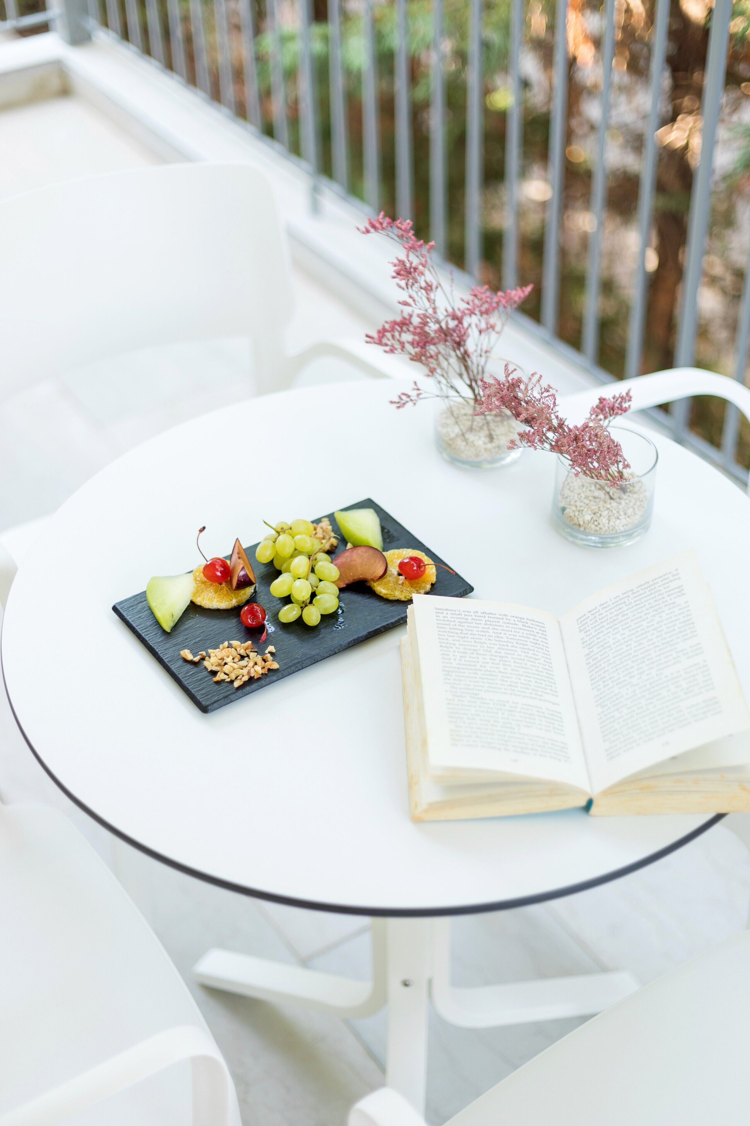 A round white table with a black slate board holding fruit and nuts, an open book, and two glass vases with dried flowers.