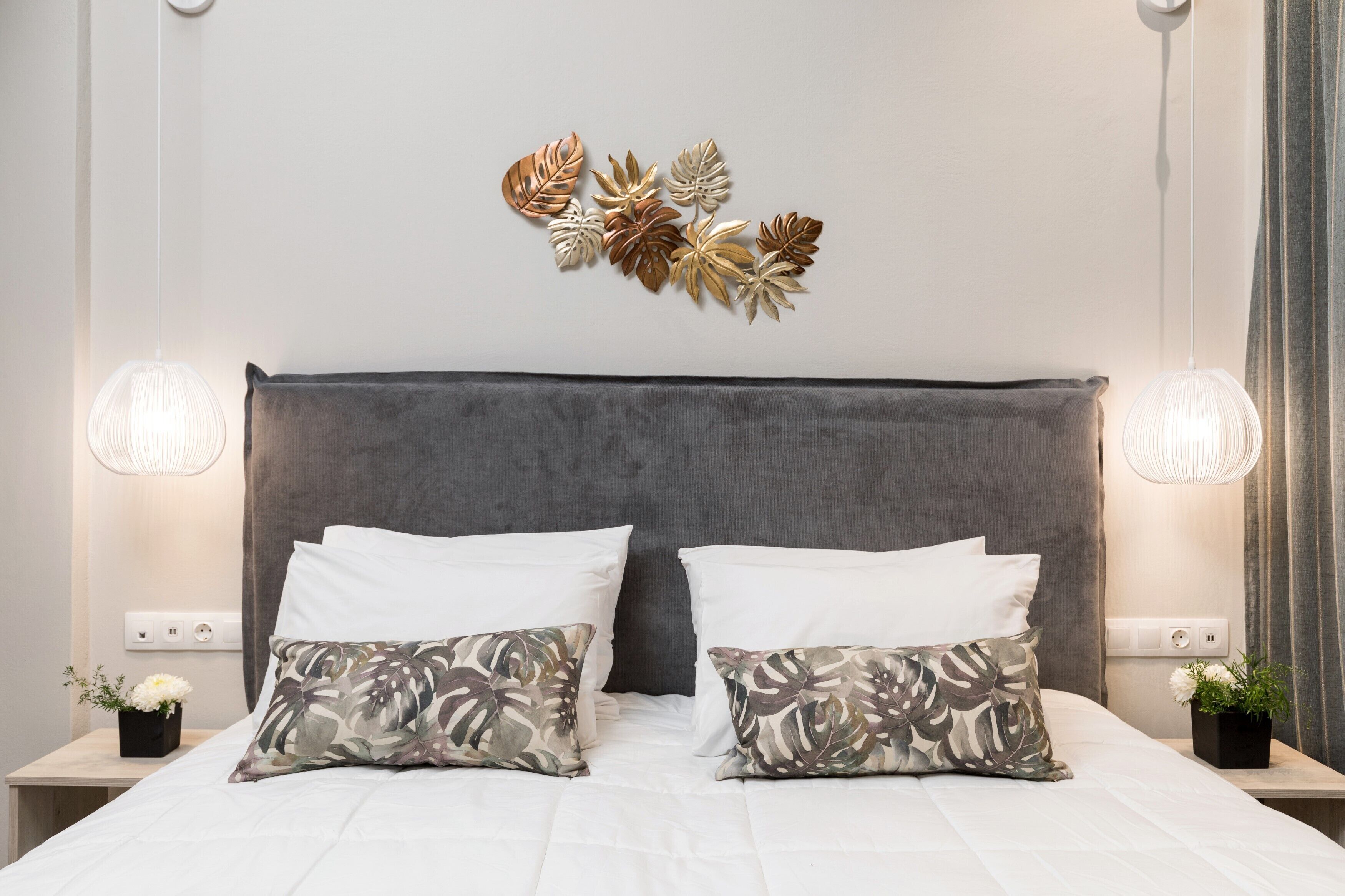 Una cama bien hecha con almohadas blancas y estampadas, un cabecero oscuro y lámparas fijadas a la pared.