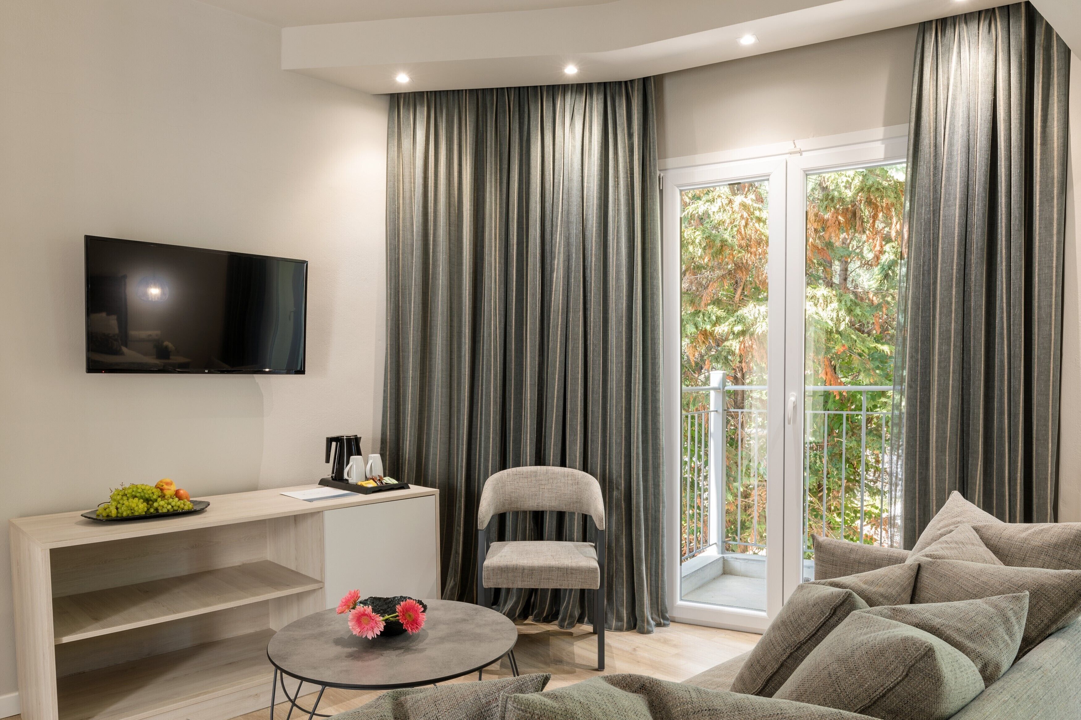 A modern living room with a flat-screen TV, a dining table with fruit, a grey armchair, and a sofa.