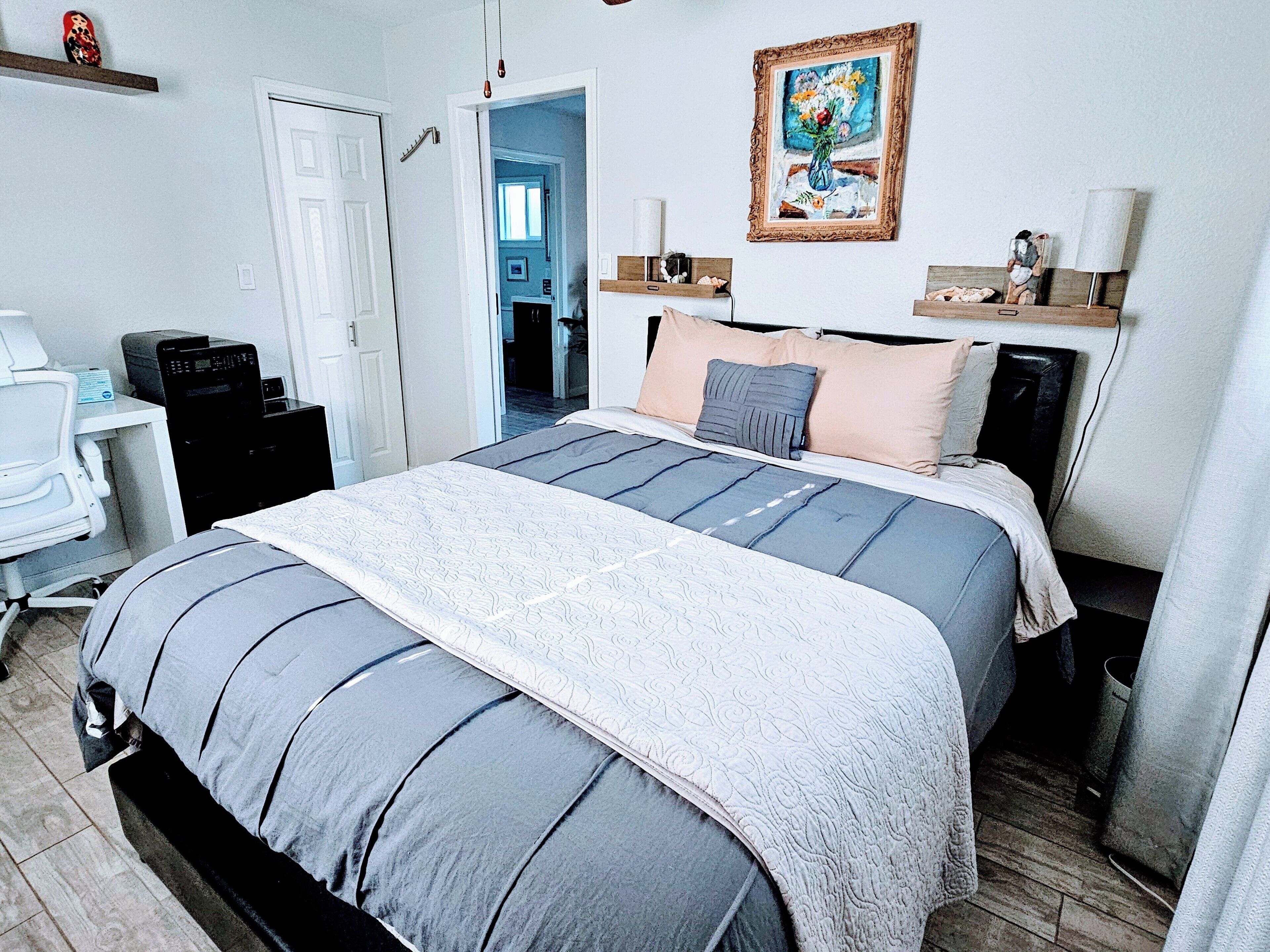 A bedroom with a bed, bedside tables, a desk, and a chair. There is a door leading to another room.