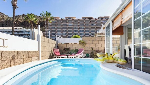 A swimming pool with lounge chairs and inflatable toys, surrounded by a stone wall and a building with large windows.