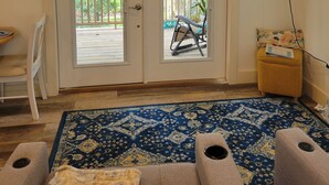 Un salon avec un tapis à motifs bleus, un téléviseur fixé au mur et une porte-fenêtre coulissante donnant sur un espace extérieur équipé d’une chaise et d’une table.