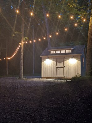 A small wooden shed with a sloped roof and a door, surrounded by trees and illuminated by string lights.