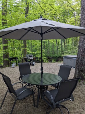 A round table with four chairs and a large umbrella in a forested area.