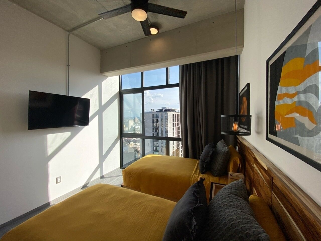 Habitación de hotel con dos camas, televisión de pantalla plana, ventana grande con vista a la ciudad y ventilador de techo.