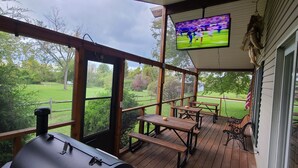 A covered outdoor deck with wooden tables and benches, a barbecue grill, and a television displaying a sports game.
