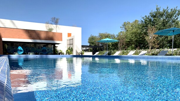 Una piscina azul cristalina con borde de azulejos, rodeada de sillas de descanso y sombrillas, y un edificio de diseño moderno al fondo.