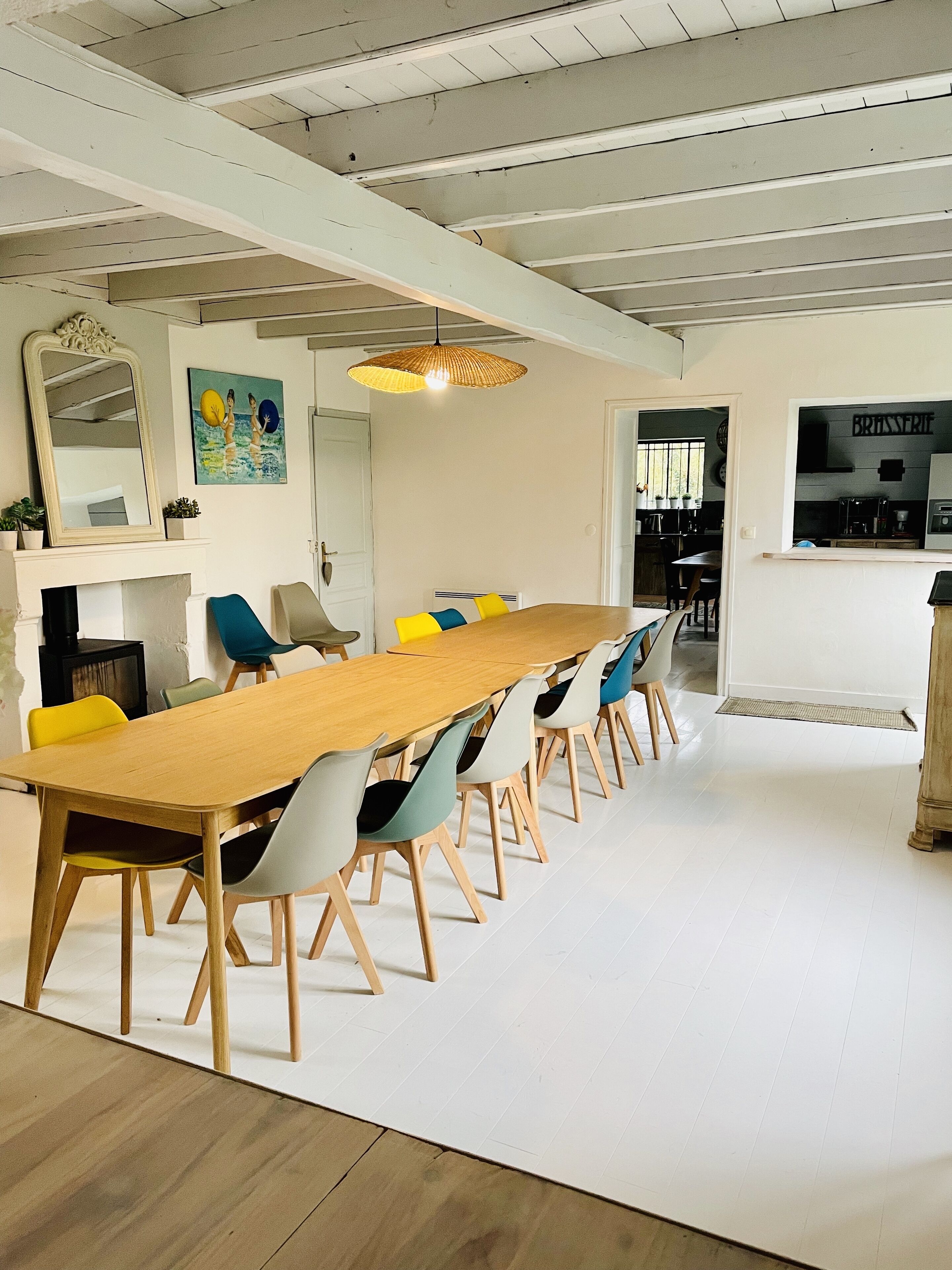 Een eethoek met een lange houten tafel, kleurrijke stoelen, een hanglamp en een zichtbare keuken door een open deur.