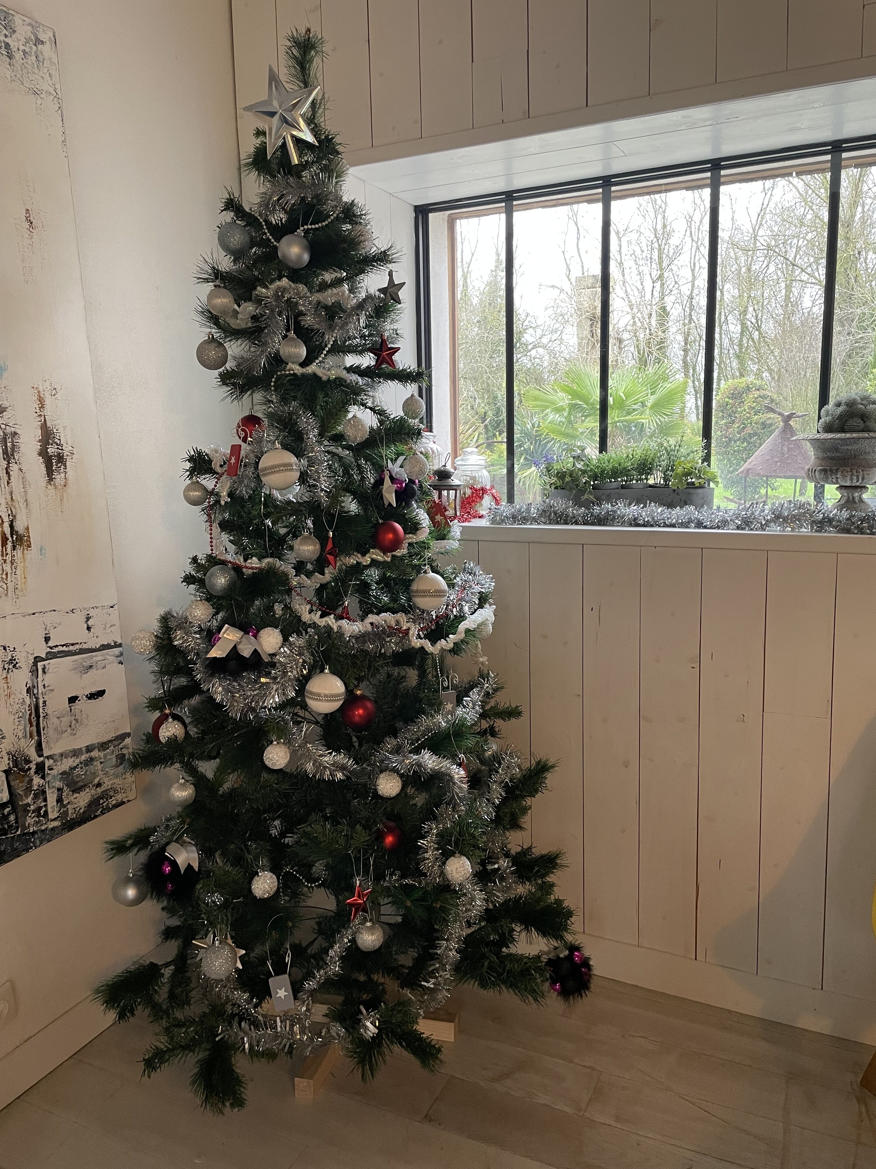 Un árbol de Navidad con adornos plateados y rojos en una habitación con paredes de madera y una ventana que da a la vegetación.