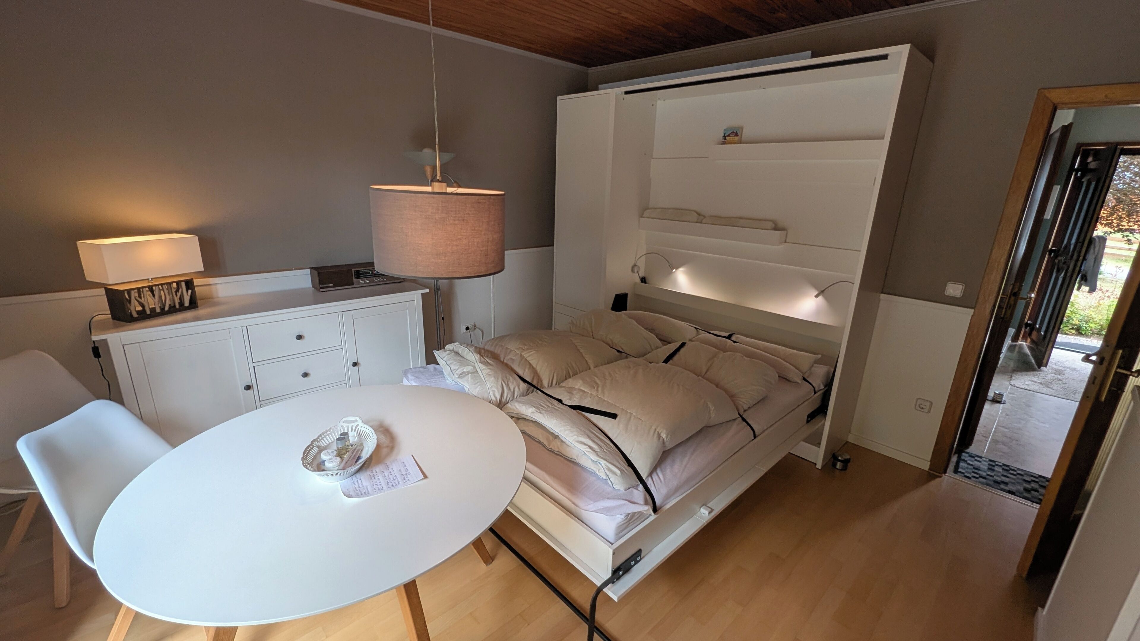 A compact room with a bed, a table, a chair, a desk with a lamp, and a hanging light fixture.