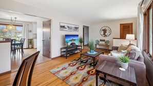 A modern living room with a sofa, coffee table, and TV. There is a dining area with a table and chairs. The room has wooden flooring and a colorful rug. - Brand New WATERFRONT Listing! (Bloomington)