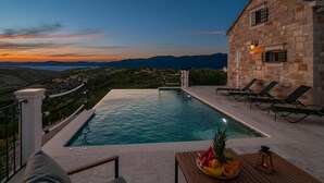 A swimming pool with a view of mountains at sunset, a stone building with a balcony, and lounge chairs. - Beautiful home in Postira (Postira)