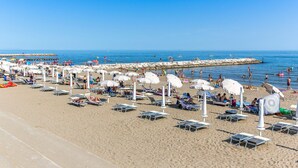 Een strand met rijen ligbedden en parasols, een rotsenrij en mensen die zwemmen en zonnen.