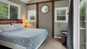 Ein Schlafzimmer mit einem Bett, einem Deckenventilator, zwei Fenstern, einer Lampe und Blick auf Bäume.