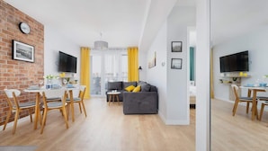 A modern living room with a grey sofa, wooden dining table, and chairs. There is a TV mounted on the wall and a window with yellow curtains. - Apartment with Balcony | Pool, Spa and Jacuzzi, 100 m to the Beach (Rewal)