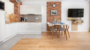 A modern kitchen with white cabinets, a brick accent wall, a dining area with a table and chairs, and a flat-screen TV mounted on the wall. - Apartment with Balcony | Pool, Spa and Jacuzzi, 100 m to the Beach (Rewal)