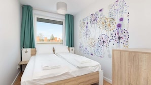 A bedroom with a wooden bed, a nightstand, a dresser, a window with curtains, and a wall with a floral pattern. - Apartment with Balcony | Pool, Spa and Jacuzzi, 100 m to the Beach (Rewal)