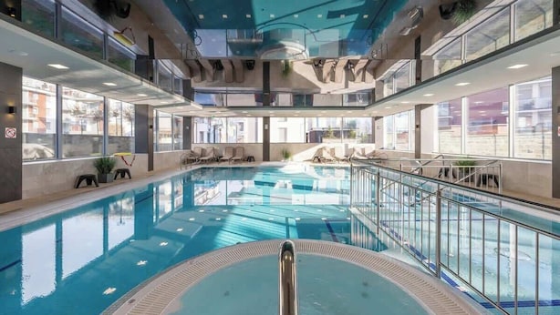 A large indoor swimming pool with clear glass walls, a central diving platform, and multiple seating areas along the sides. - Apartment with Balcony | Pool, Spa and Jacuzzi, 100 m to the Beach (Rewal)