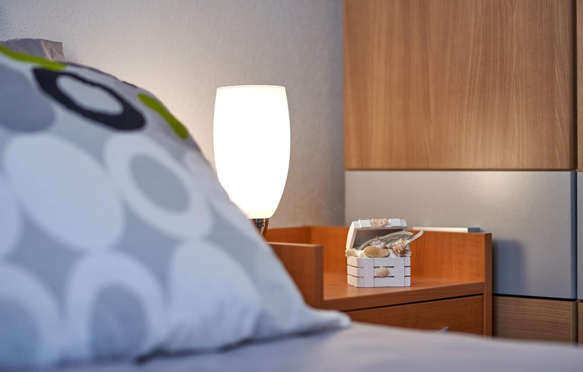 Un lit bien fait, avec un oreiller blanc orné d’un motif circulaire. Une table de chevet porte une petite boîte blanche contenant des coquillages. Une lampe éclaire la pièce.