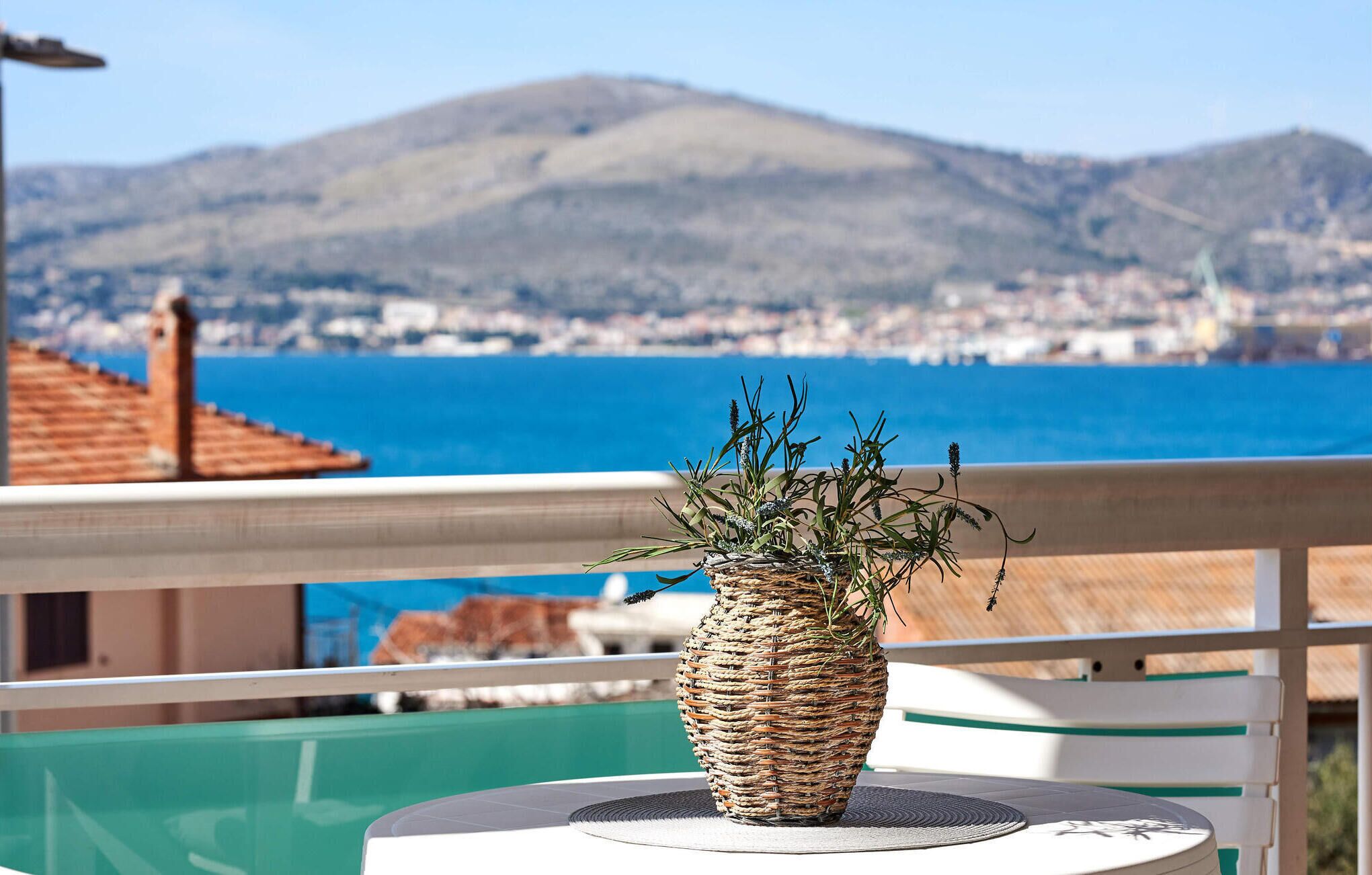 Ein Korbbesen-Vase mit Pflanzen auf einem Balkon mit Blick auf eine Küstenstadt, im Hintergrund ein Berg.