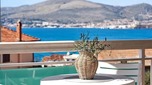Un vase en osier contenant des plantes, placé sur un balcon donnant sur une ville côtière, avec une montagne en arrière-plan.