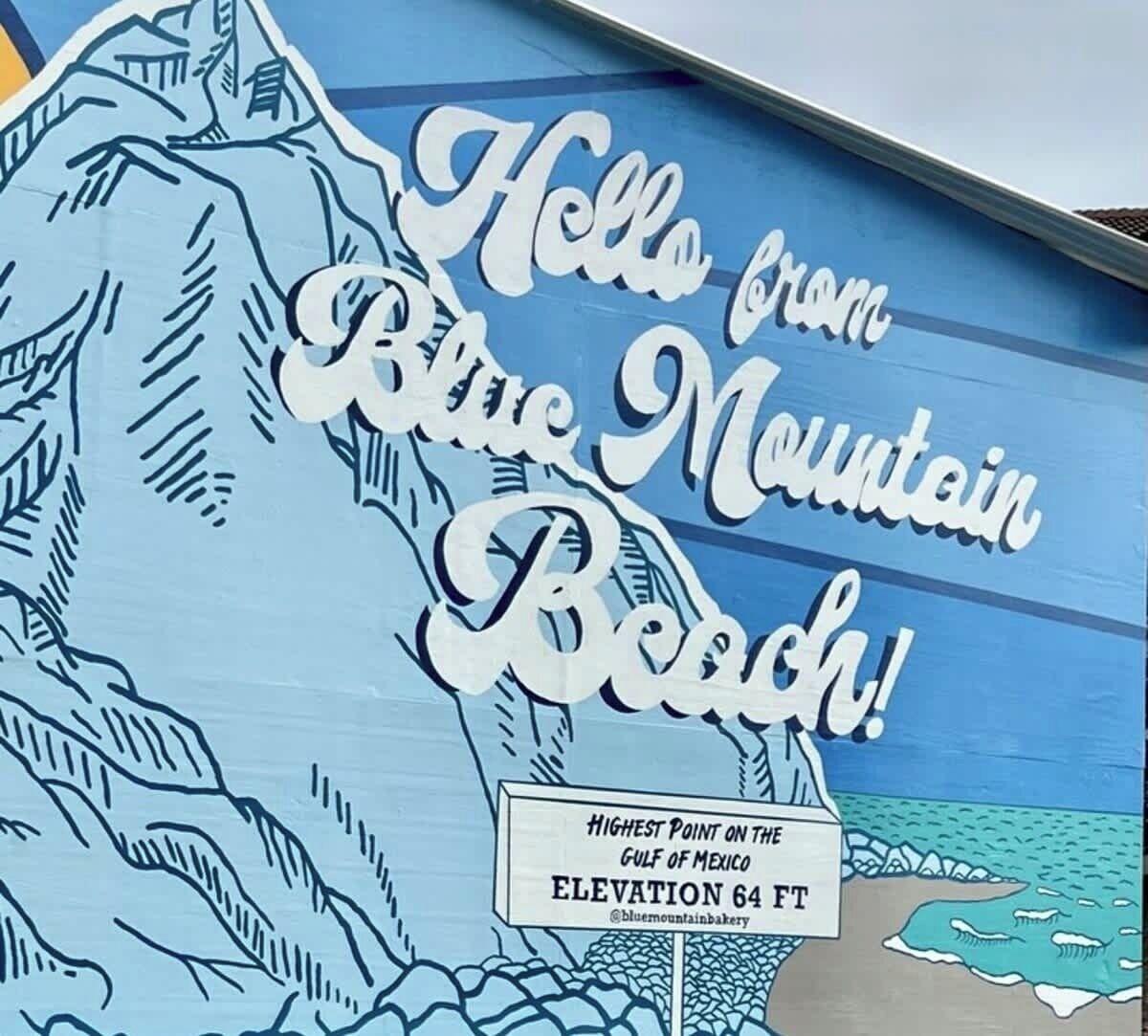 A sign with 'Hello from Blue Mountain Beach!' and 'Highest Point on the Gulf of Mexico Elevation 64 FT' text.