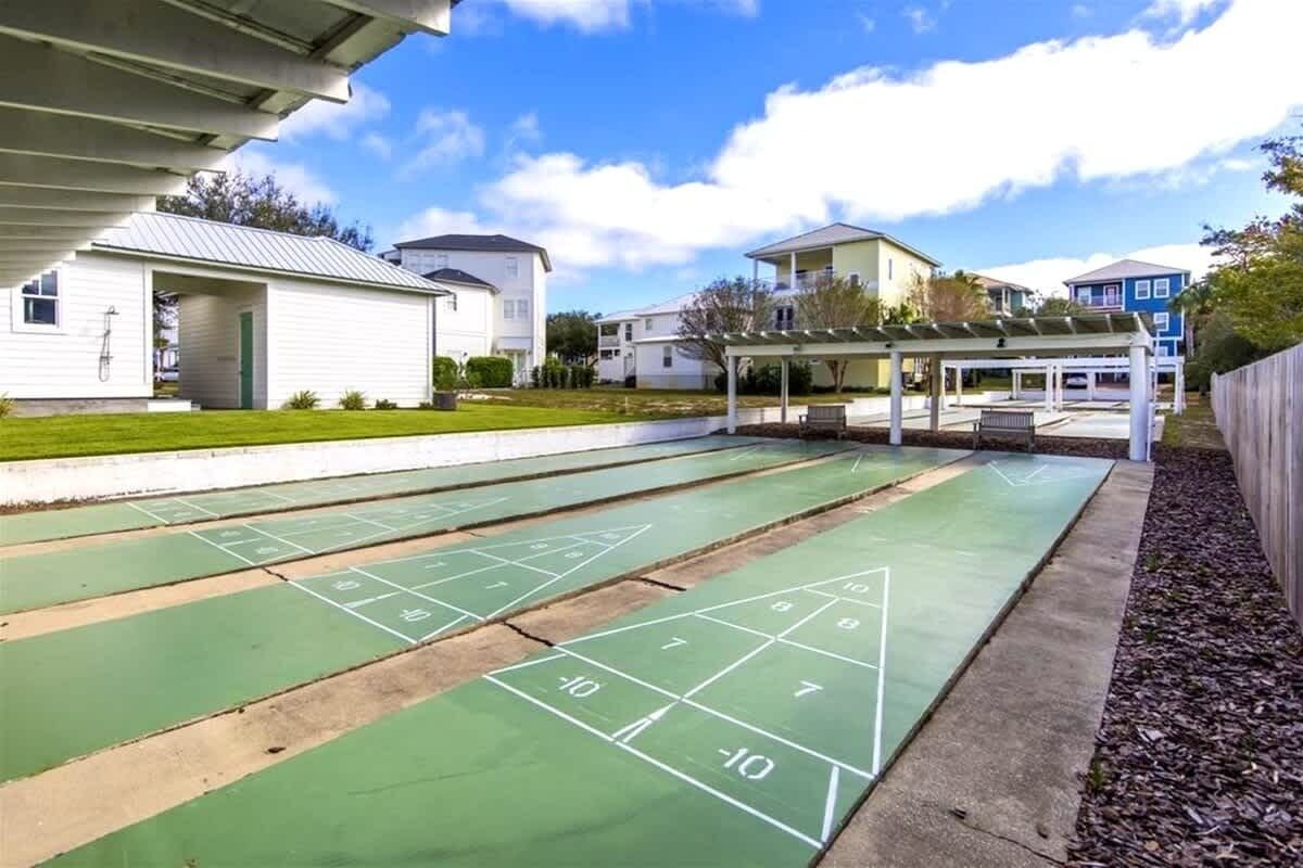 Un terrain de jeu de shuffleboard vert, avec des chiffres et des lignes, un bâtiment blanc avec une porte, et un espace abrité avec des bancs.