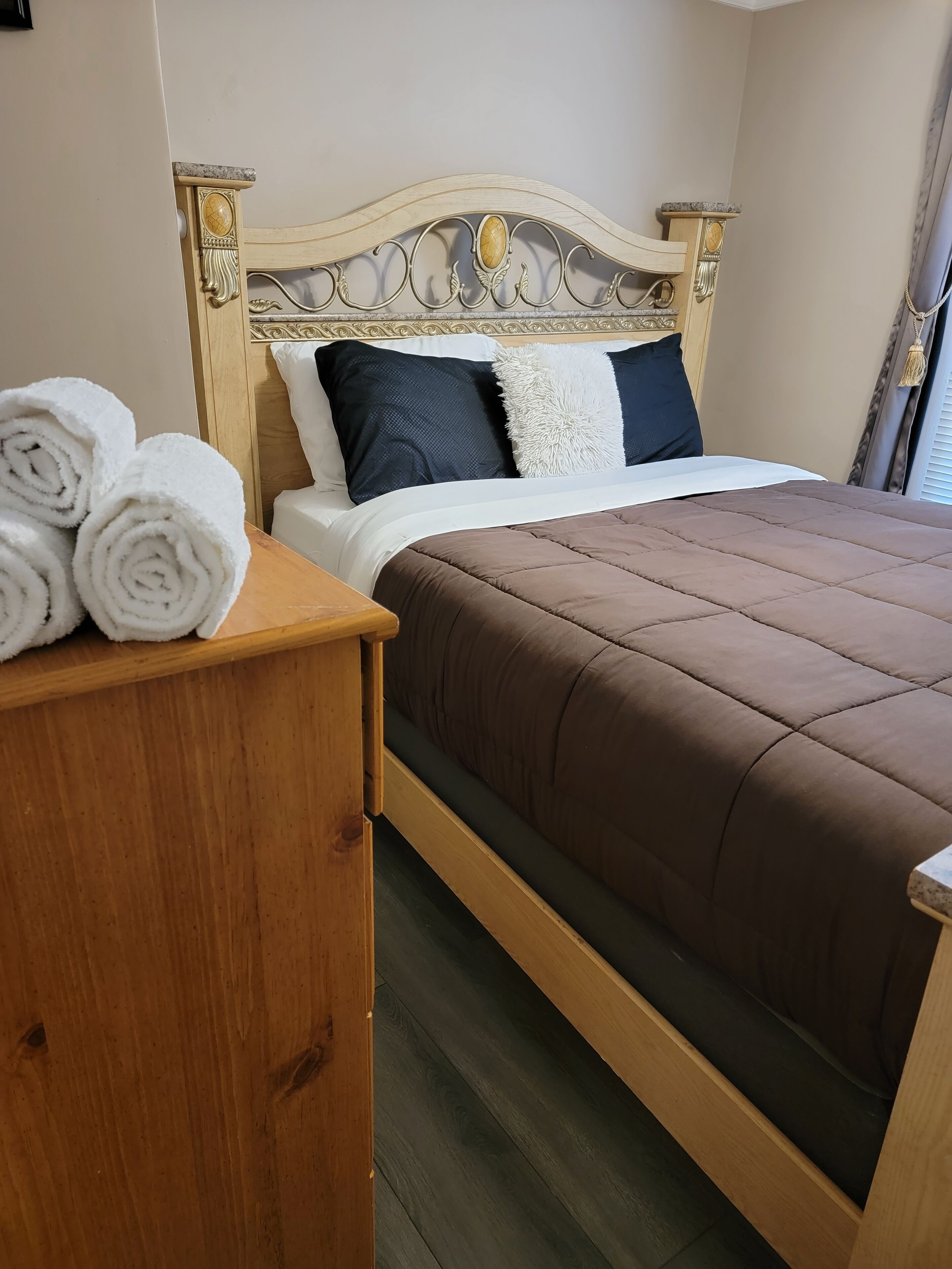 A neatly made bed with a brown quilt, a wooden headboard with decorative carvings, and rolled white towels on a wooden nightstand.