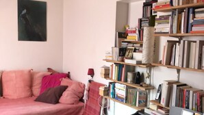 Ein gemütliches Wohnzimmer mit einem rosafarbenen Sofa, einer türkisblauen Aufbewahrungsbox, einem schwarzen Schreibtisch und einem Bücherregal voller Bücher.
