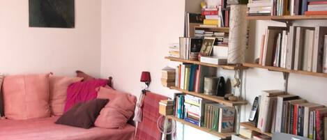 A cozy living room with a pink sofa, a teal storage box, a black desk, and a bookshelf filled with books.