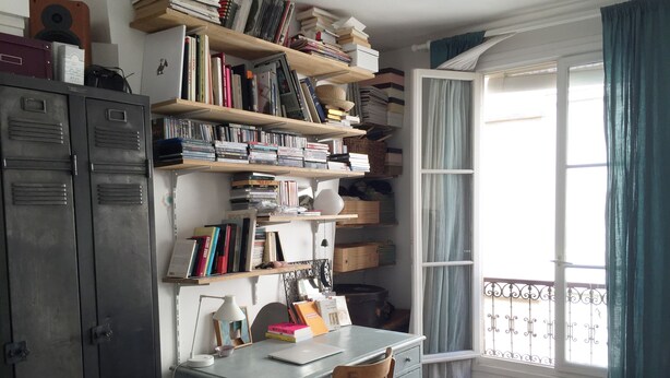 Ein Arbeitsraum mit Schreibtisch, Regalen mit Büchern, einem Stuhl und einem Fenster mit Vorhängen.