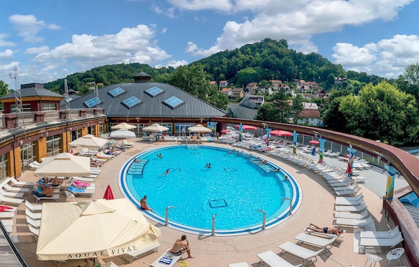 A circular pool with lounge chairs and umbrellas, surrounded by a building with a sloped roof and a view of a forested hill.