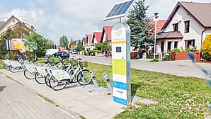 A row of bicycles parked on a sidewalk, a solar panel on a pole, and houses in the background.