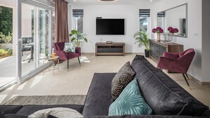 Una sala de estar moderna con un televisor de pantalla plana grande, un sofá con cojines estampados, un sillón rojo y una zona de comedor con una mesa de vidrio y sillas.