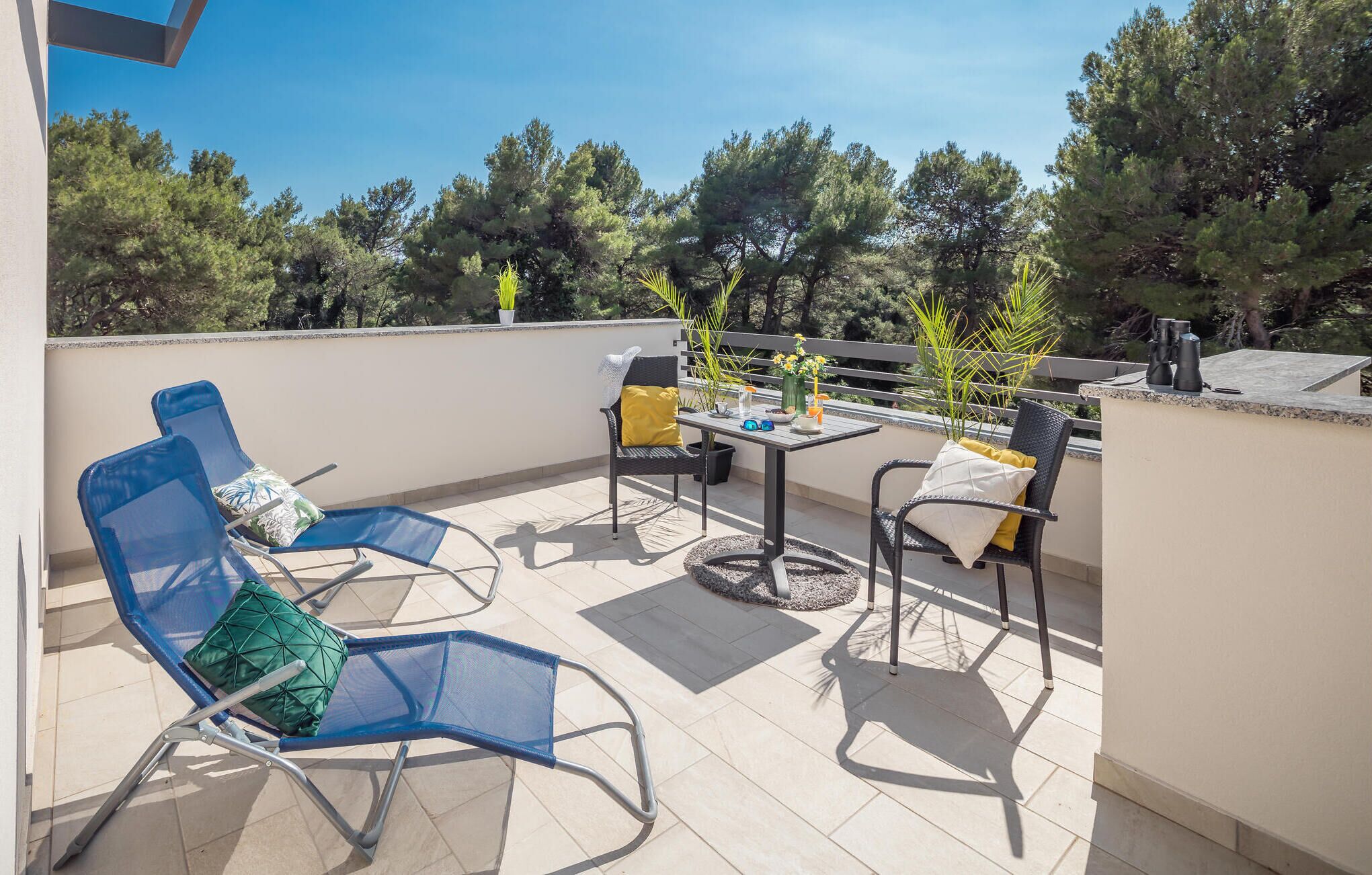 Een balkon met twee blauwe stoelen, een kleine tafel met een bloevas, en een potplant.